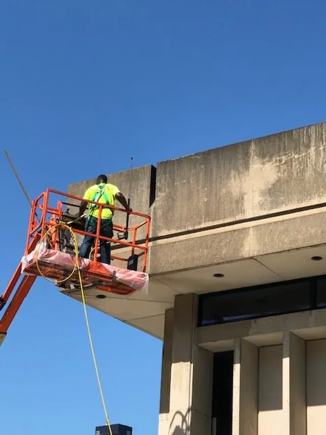 Crew member in an orange lift basket cleaning the concrete fascia of a building under blue sky