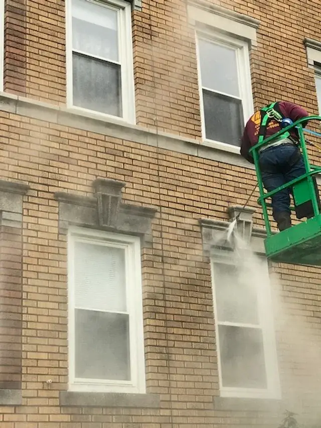 Crew member in a lift basket washing a tan brick facade beside a window, spray in the air