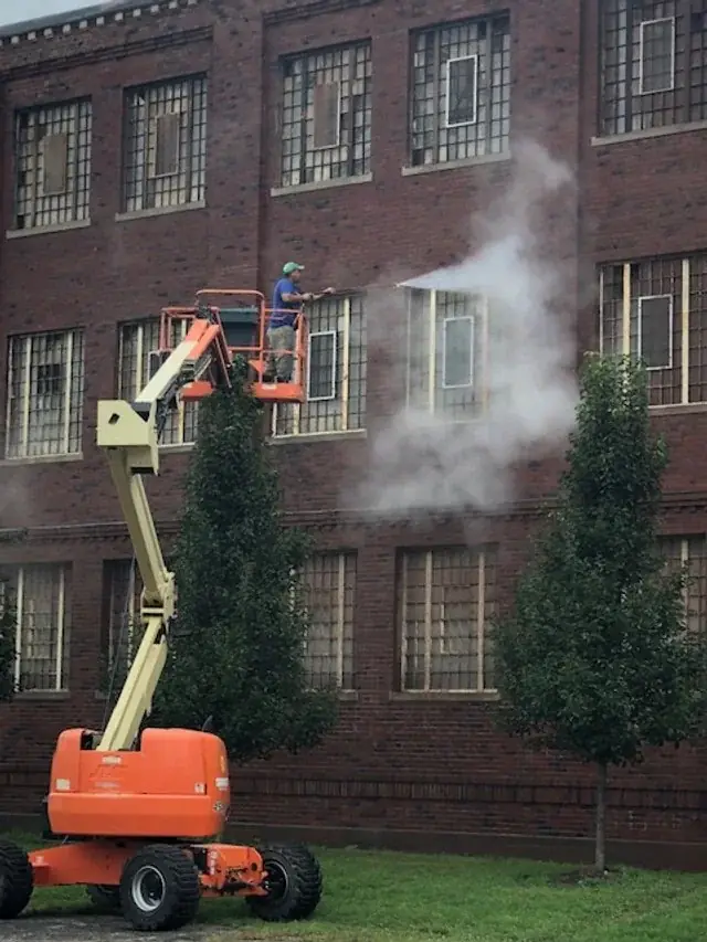 Orange boom lift raised against a dark brick building with steam rising off the facade